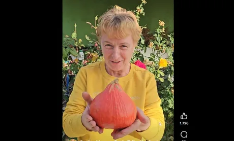 O femeie zâmbitoare ține un dovleac Hokkaido în grădină, înconjurată de flori.
