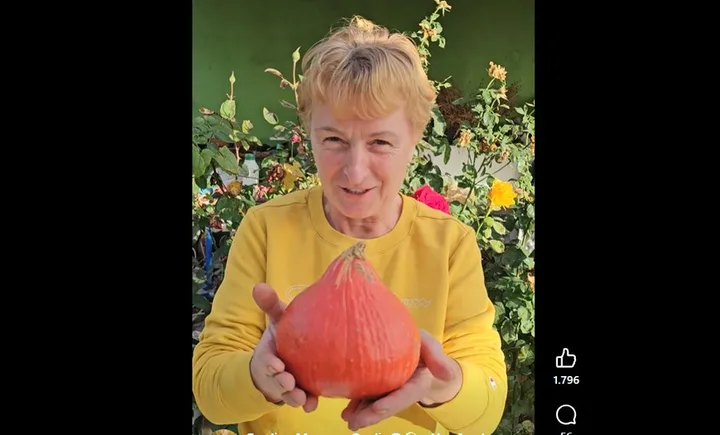 O femeie zâmbitoare ține un dovleac Hokkaido în grădină, înconjurată de flori.