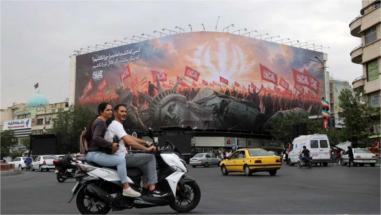 Oameni pe motocicletă în fața unui panou publicitar mare în Teheran.