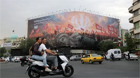 Oameni pe motocicletă în fața unui panou publicitar mare în Teheran.