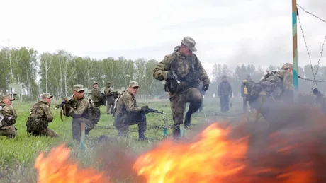 Soldați în exercițiu militar, cu foc și vegetație în fundal.
