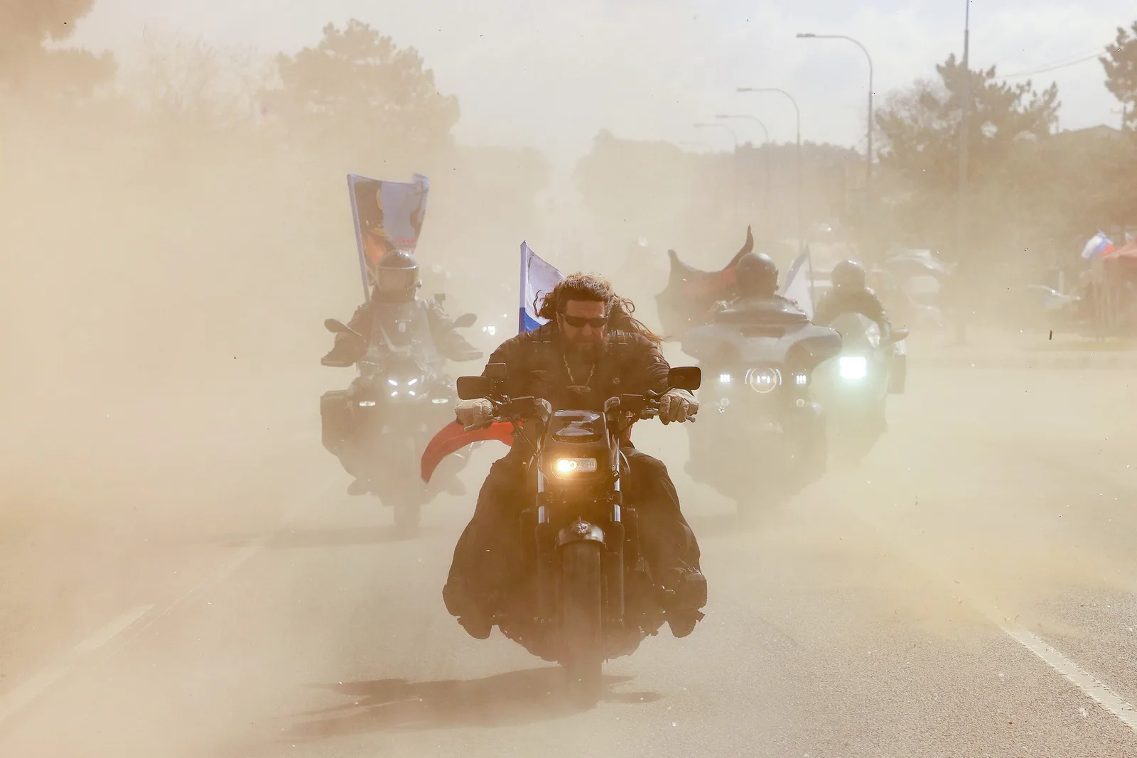 Motocicliști pe o șosea acoperită de praf, înconjurați de steaguri.