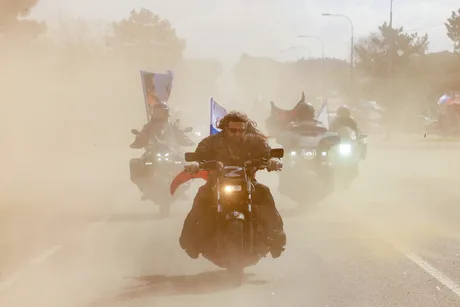 Motocicliști pe o șosea acoperită de praf, înconjurați de steaguri.