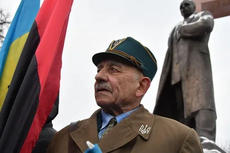 Un bărbat cu pălărie stă lângă un monument, purtând steaguri ucrainene.