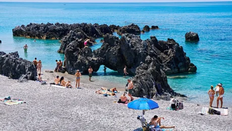 Plajă aglomerată cu oameni, stânci și umbrele colorate pe nisip fin.