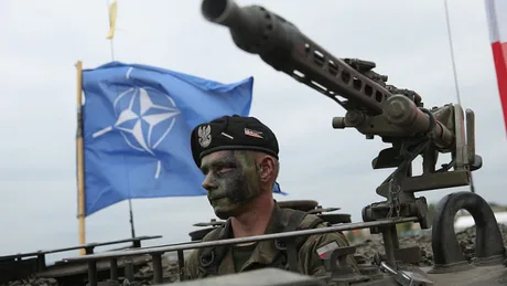 Soldat NATO în fața unui steag, pregătit pentru misiune militară.