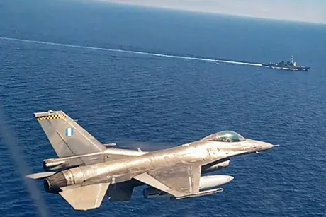 Avion de vânătoare F-16 în zbor deasupra mării, cu un vas militar în fundal.
