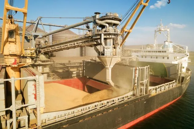 Încărcare de grâu pe un vas în port, cu echipamente de descărcare vizibile.