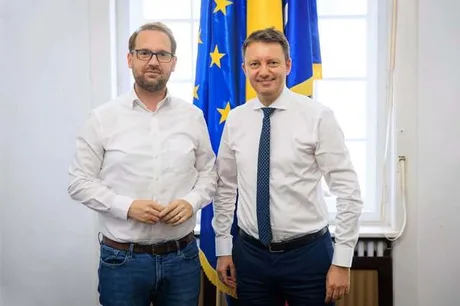 Dominic Fritz și un coleg în fața drapelului Uniunii Europene.