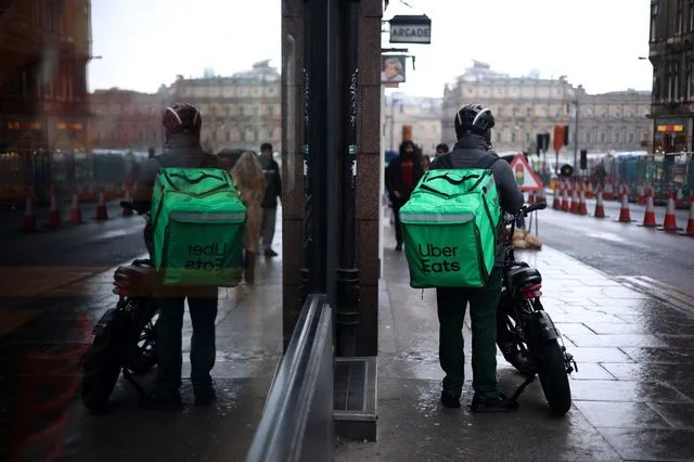 Curier Uber Eats așteptând pe o stradă aglomerată, cu rucsacul verde pe spate.