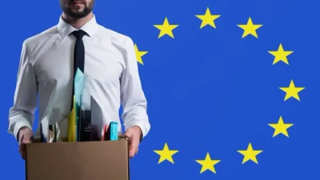 Un bărbat îmbrăcat la costum ține o cutie cu obiecte, pe fundalul steagului Uniunii Europene.