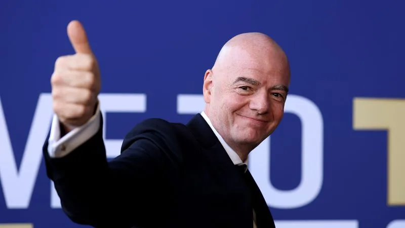 Gianni Infantino zâmbind și făcând semnul thumbs-up în fața unui panou FIFA.