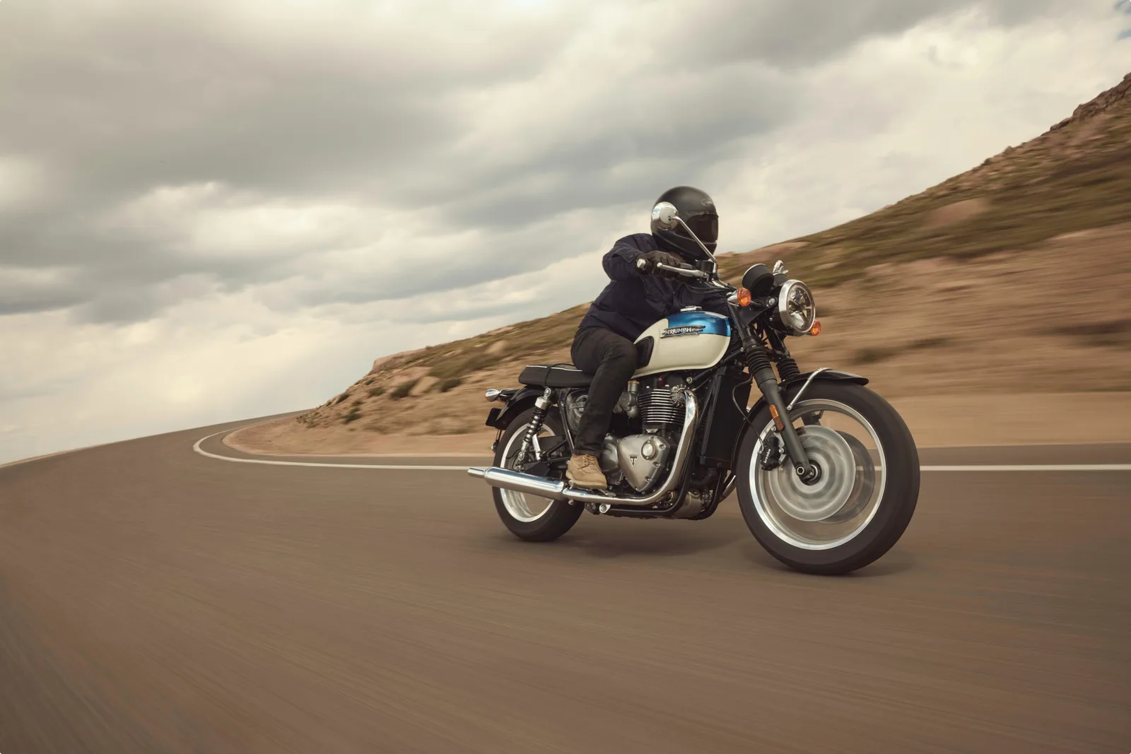 Motocicletă Triumph pe drum, cu rider echipat, peisaj montan în fundal.