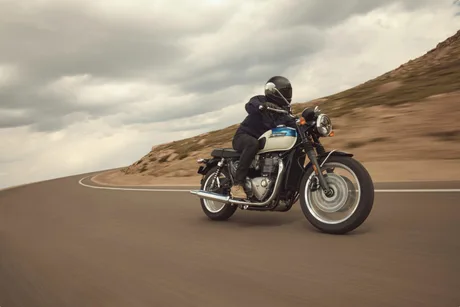 Motocicletă Triumph pe drum, cu rider echipat, peisaj montan în fundal.
