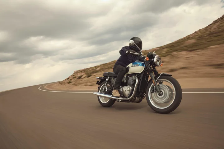 Motocicletă Triumph pe drum, cu rider echipat, peisaj montan în fundal.