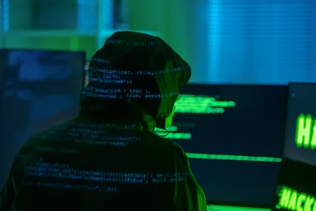 Un hacker lucrează la un computer, înconjurat de ecrane verzi.