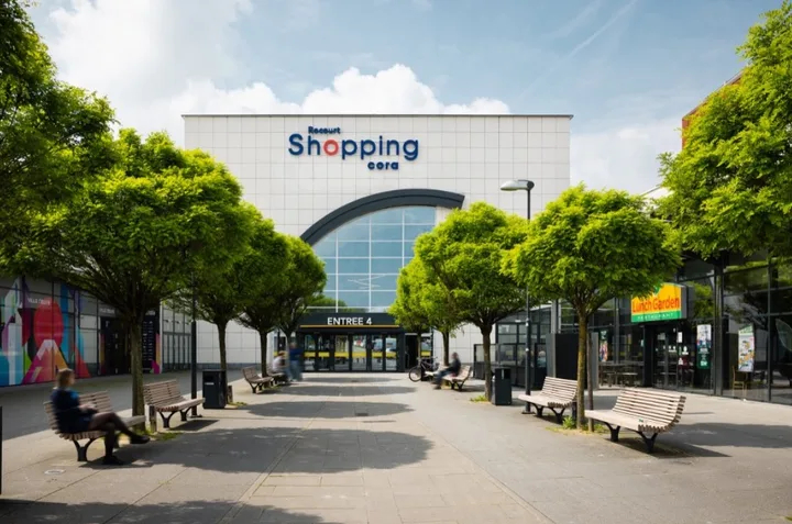 Intrarea principală a centrului comercial Shopping Cora din Belgia, cu bănci și copaci.