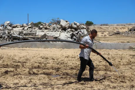 Un agricultor lucrează pe un teren afectat, cu ruine în fundal.