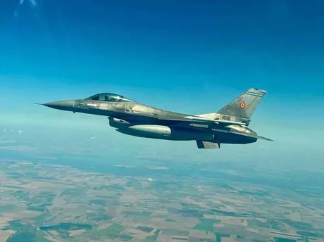 Un avion de luptă zboară deasupra unui peisaj rural vast.