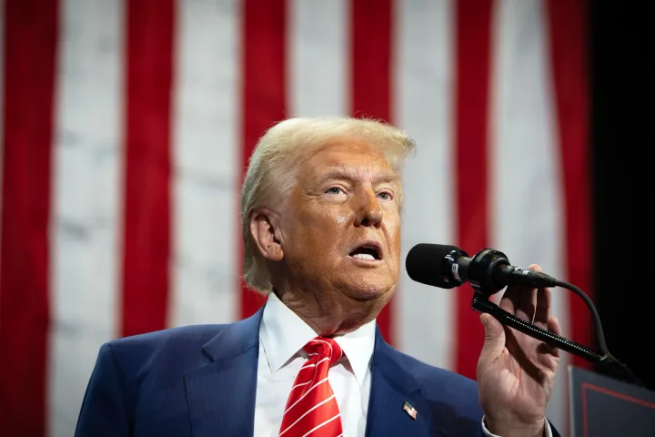 Donald Trump vorbește la microfon, cu steagul american în fundal.