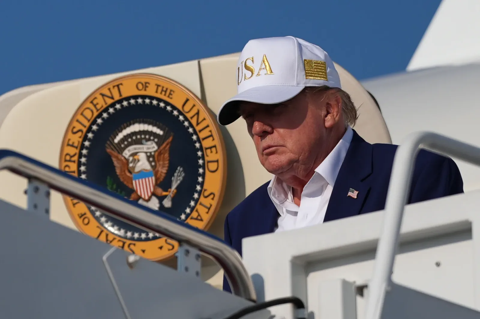 Donald Trump părăsind un avion, cu o șapcă pe cap și privirea concentrată.