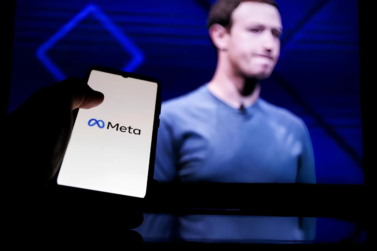 Logo-ul Meta pe un telefon, cu Mark Zuckerberg în fundal.