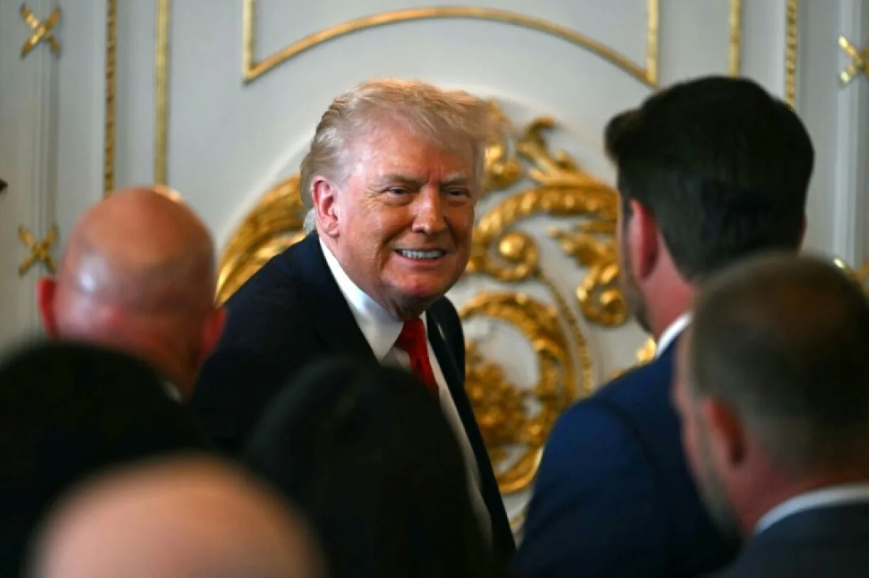 Donald Trump zâmbind în timpul unei întâlniri oficiale, înconjurat de oameni.