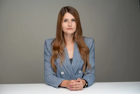 O femeie cu părul lung, îmbrăcată elegant, stă la birou cu mâinile împreunate.