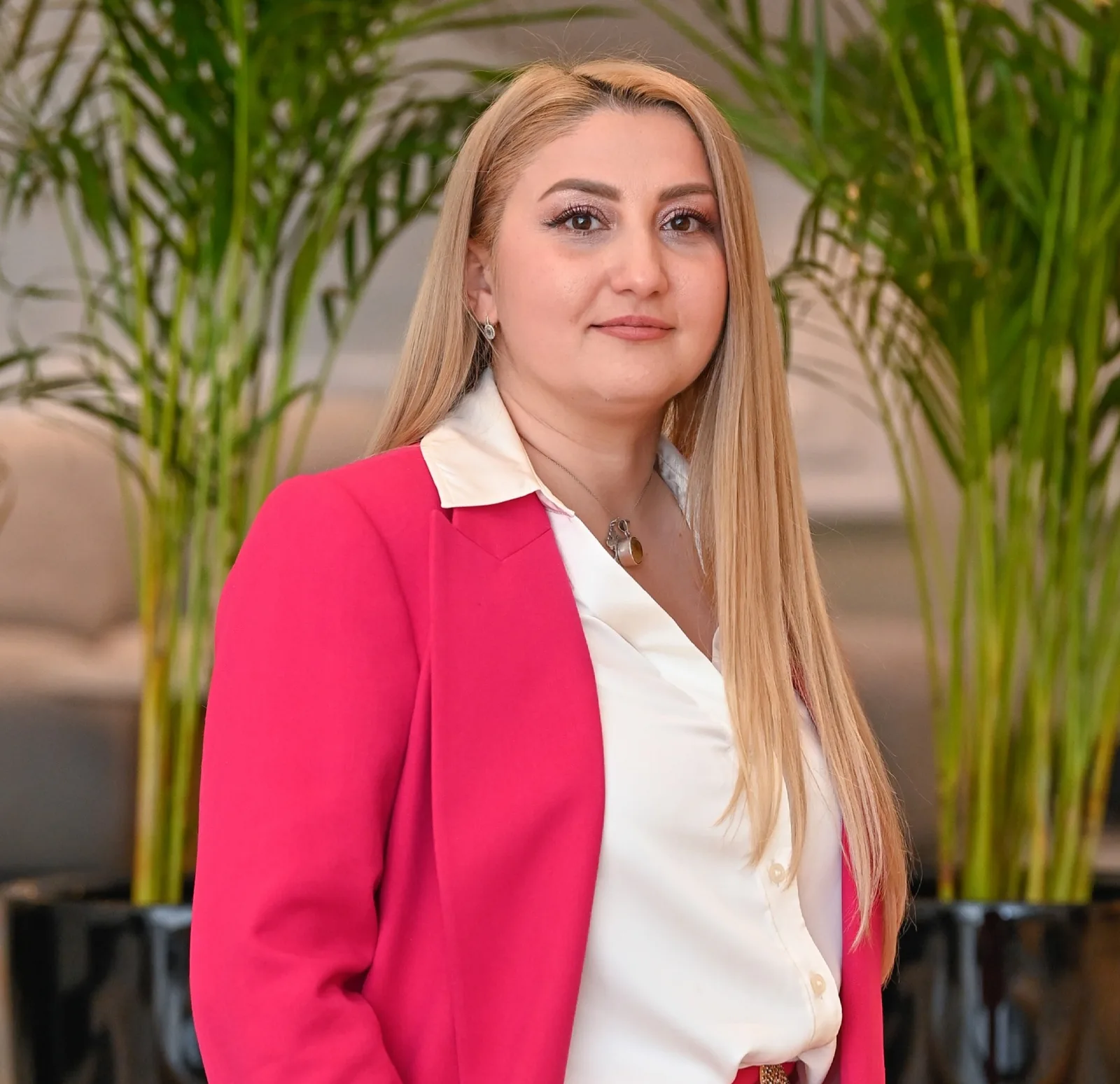 O femeie cu păr lung, îmbrăcată elegant, stând în fața unor plante verzi.