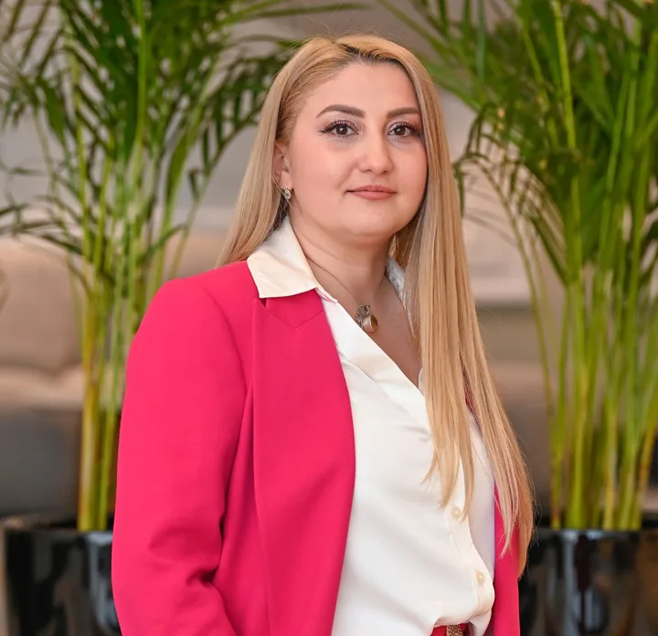 O femeie cu păr lung, îmbrăcată elegant, stând în fața unor plante verzi.