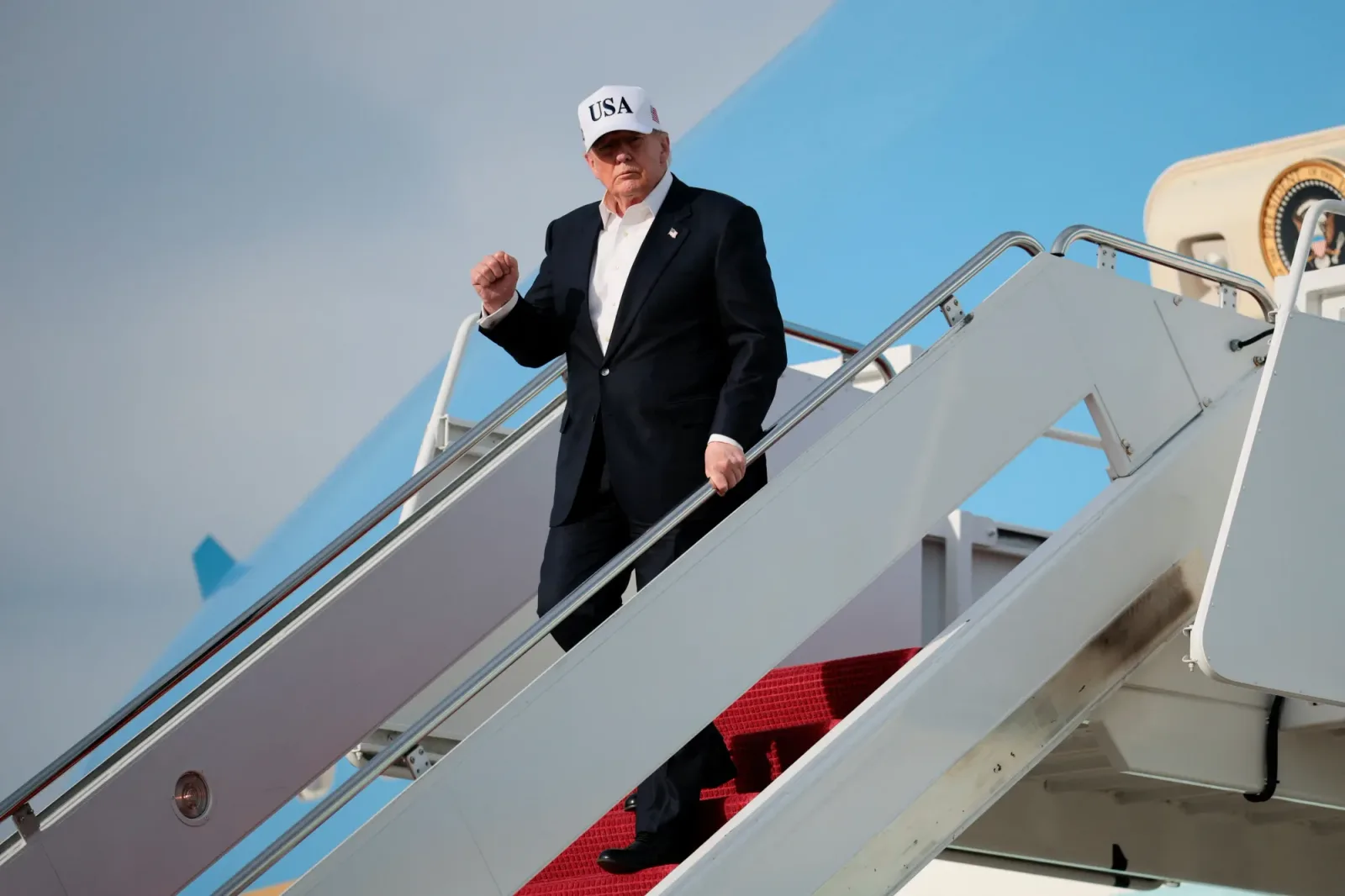 Donald Trump coborând dintr-un avion, purtând o șapcă albă cu inscripția SUA.