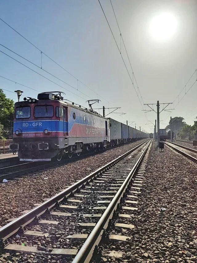 Locomotivă pe calea ferată, transportând vagoane de marfă în soare.