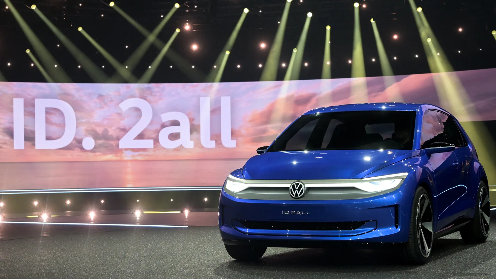 Volkswagen ID. 2all expus la un eveniment auto, cu lumini strălucitoare în fundal.