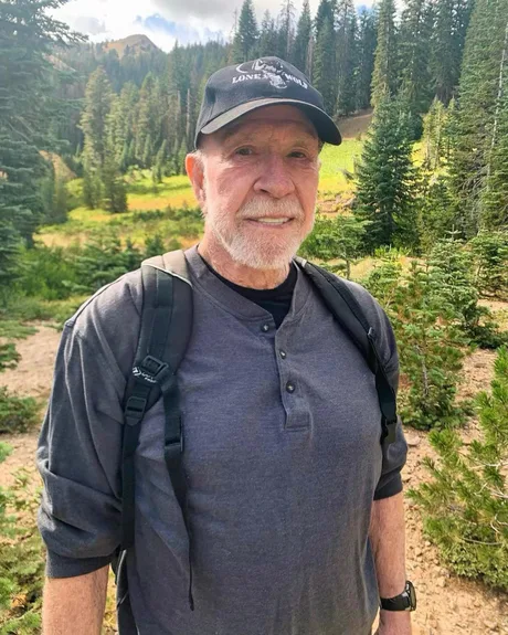 Chuck Norris zâmbind în natură, purtând o pălărie și rucsac.