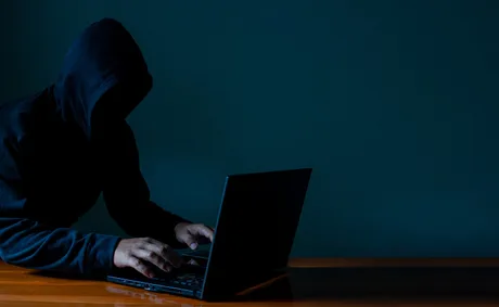 Persoană cu glugă lucrând la un laptop într-un mediu întunecat.