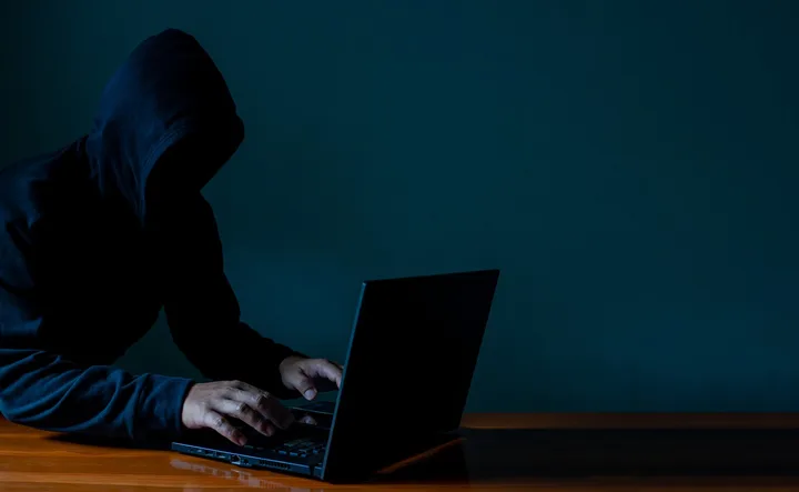 Persoană cu glugă lucrând la un laptop într-un mediu întunecat.