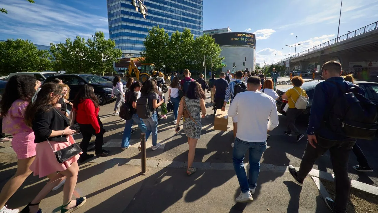 Oameni traversând o stradă aglomerată în oraș, însoțiți de clădiri moderne.
