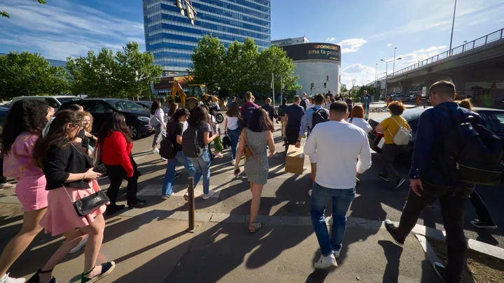 Oameni traversând o stradă aglomerată în oraș, însoțiți de clădiri moderne.