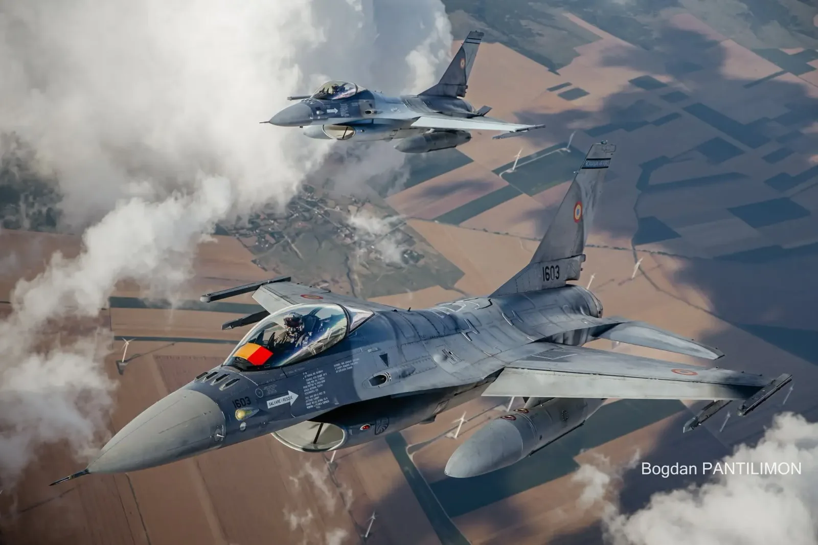 Avioane F-16 românești în zbor, pe fundal de nori și câmpuri.