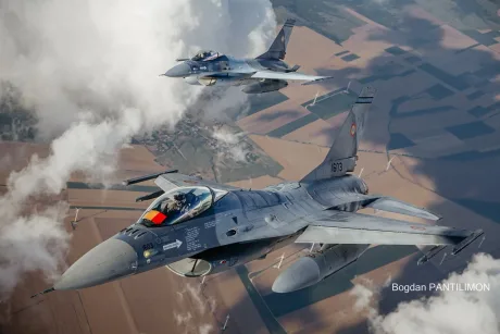 Avioane F-16 românești în zbor, pe fundal de nori și câmpuri.