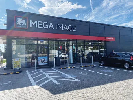 Fațada unui supermarket Mega Image, cu parcarea și clienți la intrare.