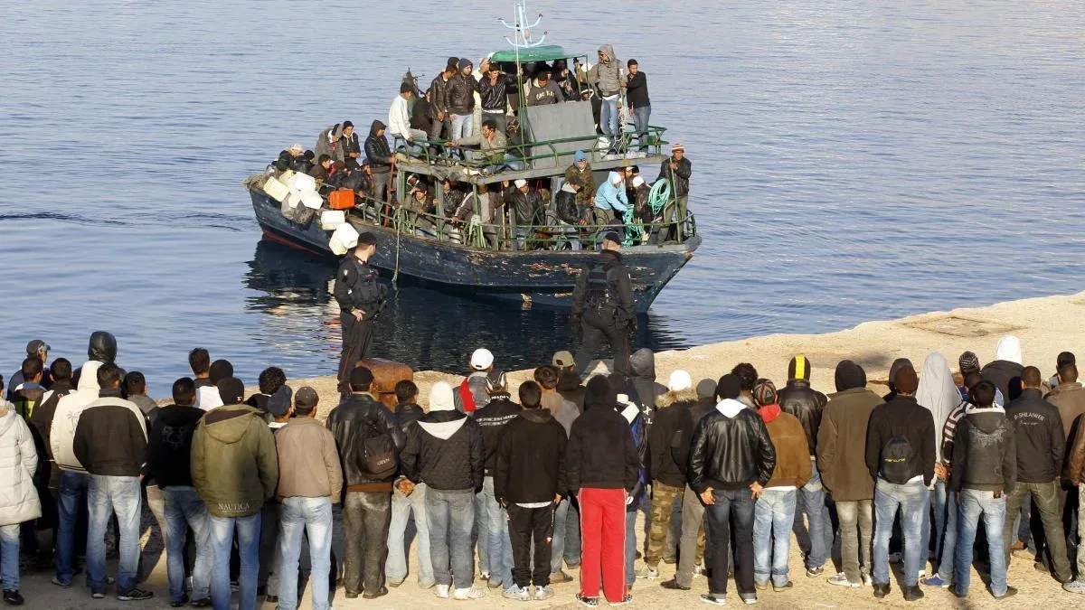 Grup de migranți pe o barcă, în fața unei plaje aglomerate.