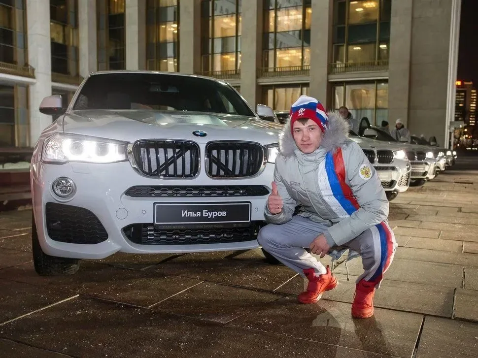 Tânăr lângă un BMW X4, cu zâmbet și degetul mare ridicat, în fața unei clădiri moderne.