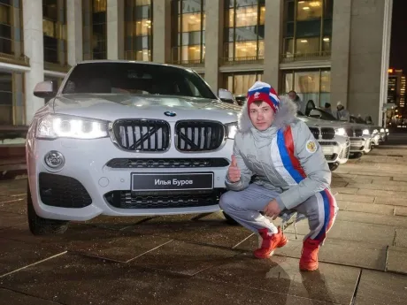 Tânăr lângă un BMW X4, cu zâmbet și degetul mare ridicat, în fața unei clădiri moderne.