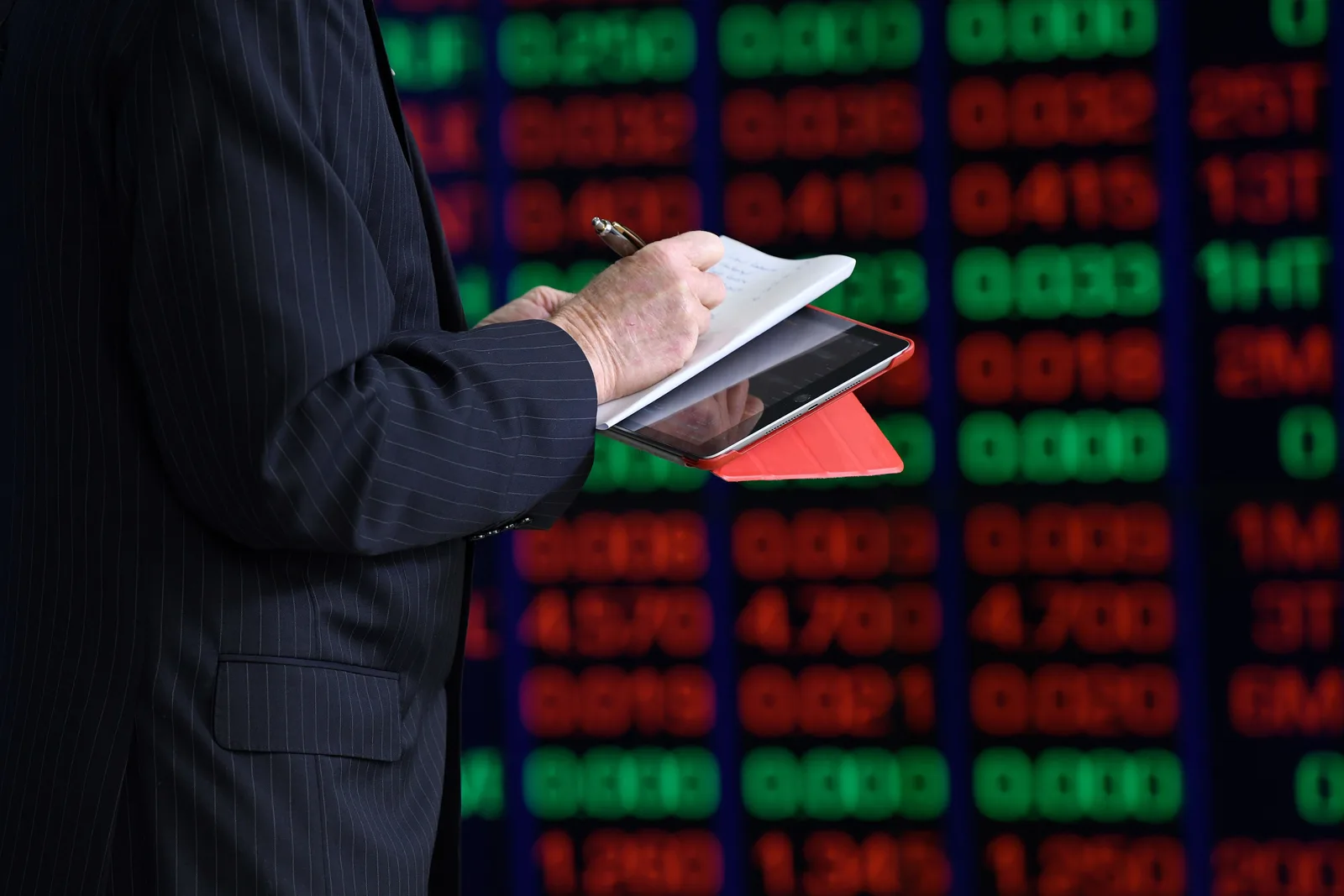 Investitor analizând date financiare pe un tabletă în fața unui panou bursier.