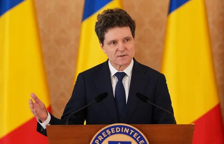Nicușor Dan discutând despre stabilitatea economică a României în fața steagurilor naționale.
