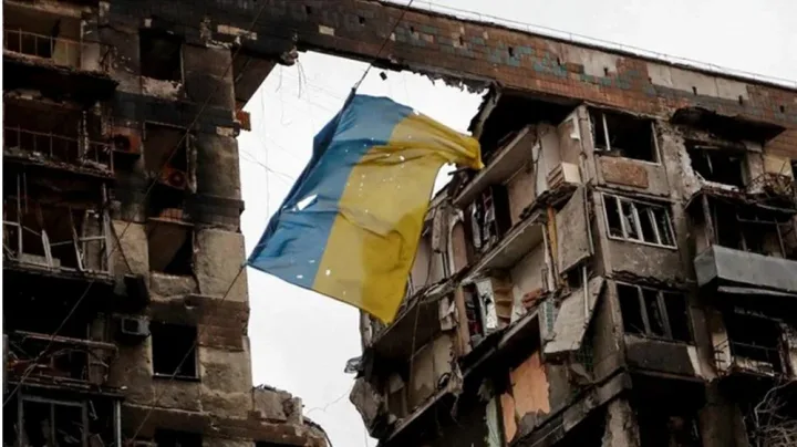Steagul Ucrainei flutură în fața unei clădiri distruse.