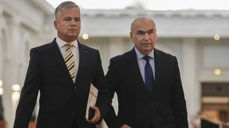 Doi bărbați în costume discutând într-un mediu formal, posibil în cadrul unei întâlniri politice.