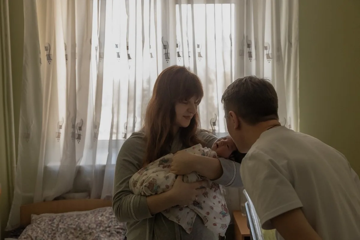 O mamă își ține copilul în brațe, zâmbind alături de tată.
