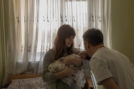 O mamă își ține copilul în brațe, zâmbind alături de tată.